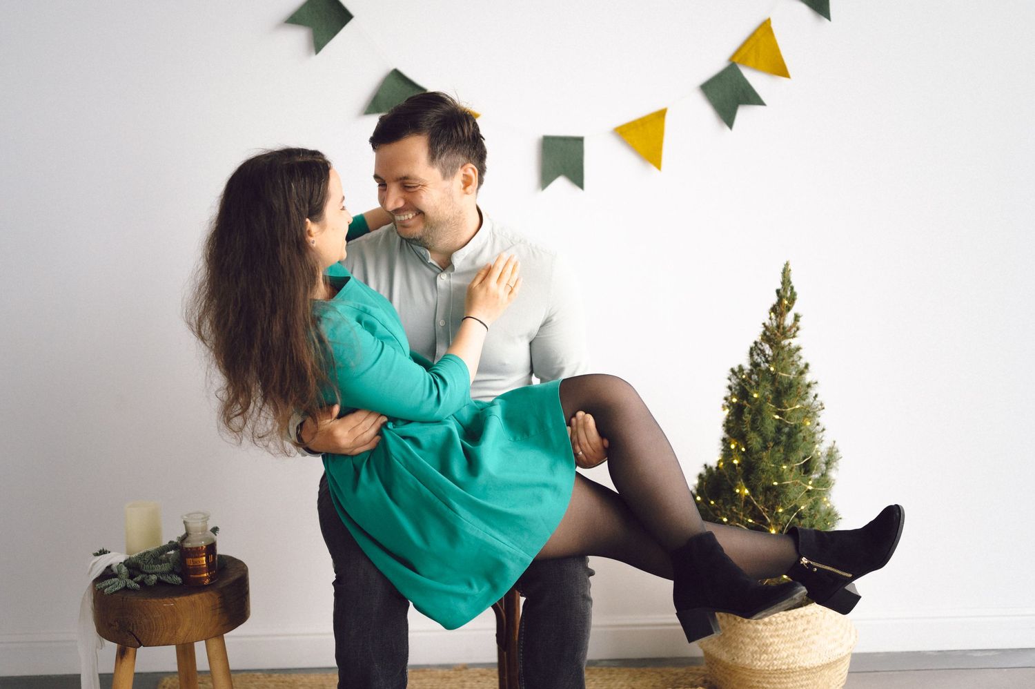 A couple in festive attire embraces in front of holiday decorations including a small Christmas tree and bunting.