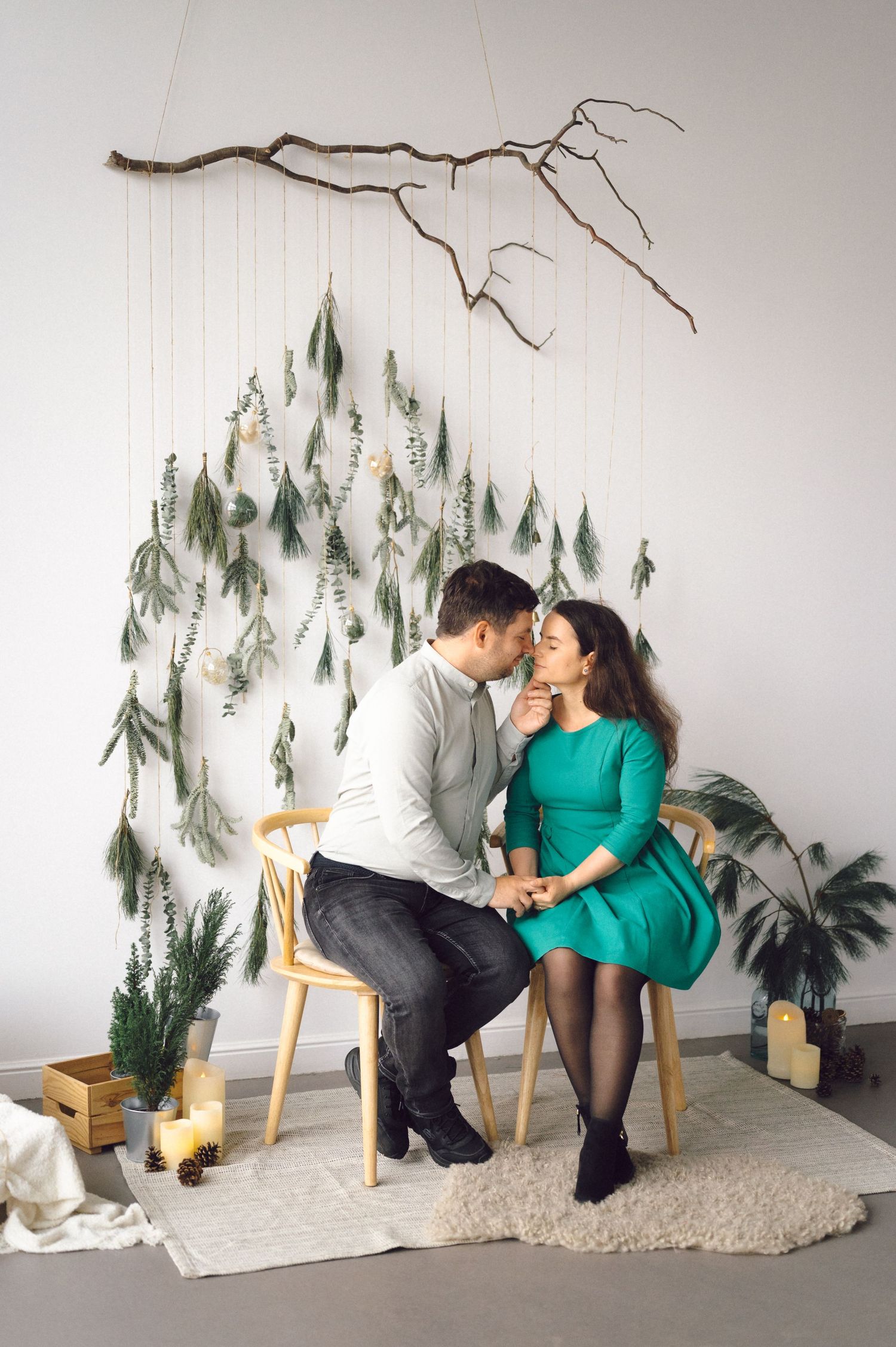 Romantic couple poses in a winter-themed photoshoot with pine trees and wooden chairs against a white backdrop.