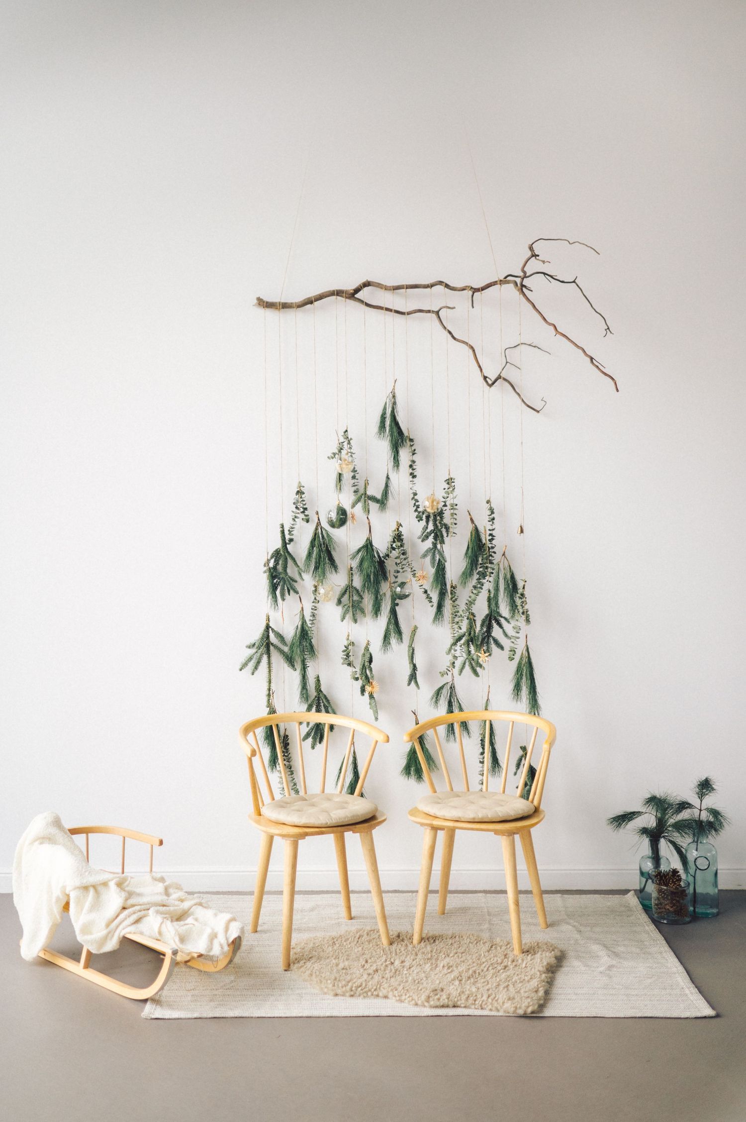 A minimalist Christmas tree installation made from hanging branches and pine boughs on mid-century modern chairs against a white wall.