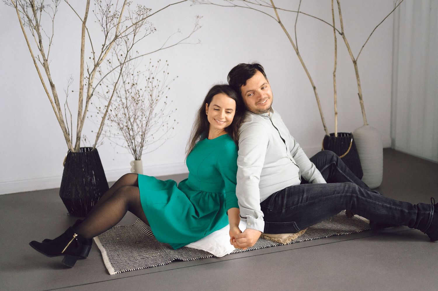 Two people in casual clothing sit together against a white wall with decorative branches.