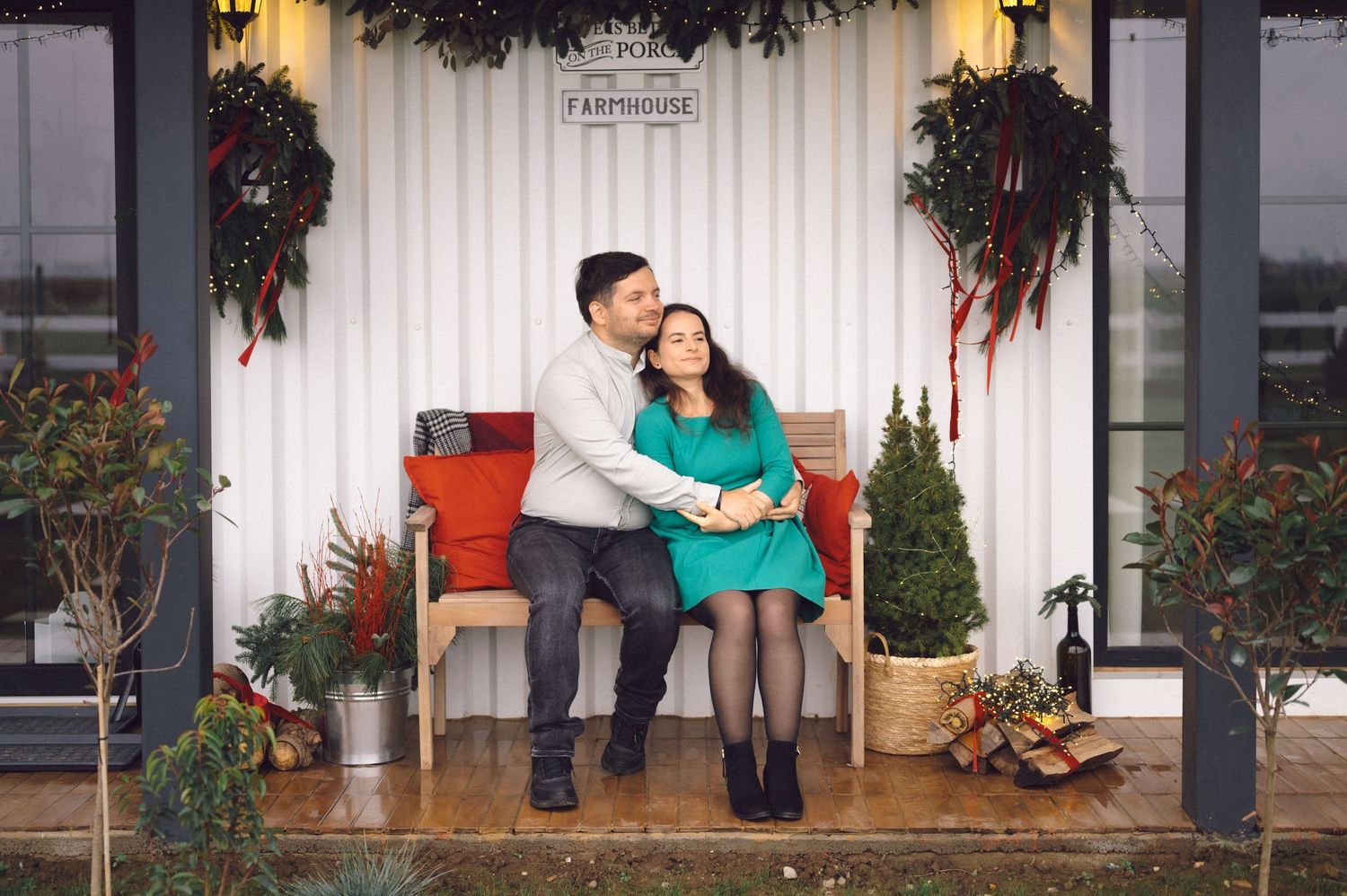 Couple in winter outfits share intimate moments on a front porch decorated with holiday greenery and wreaths.
