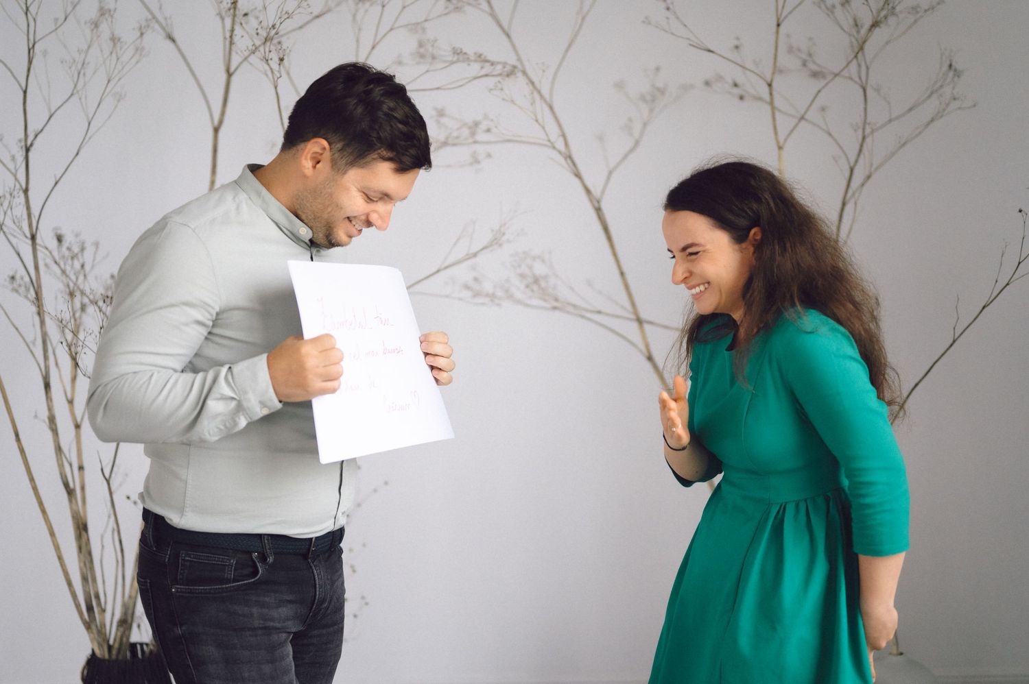 Two people in office attire stand viewing papers together against a white wall with decorative branches.