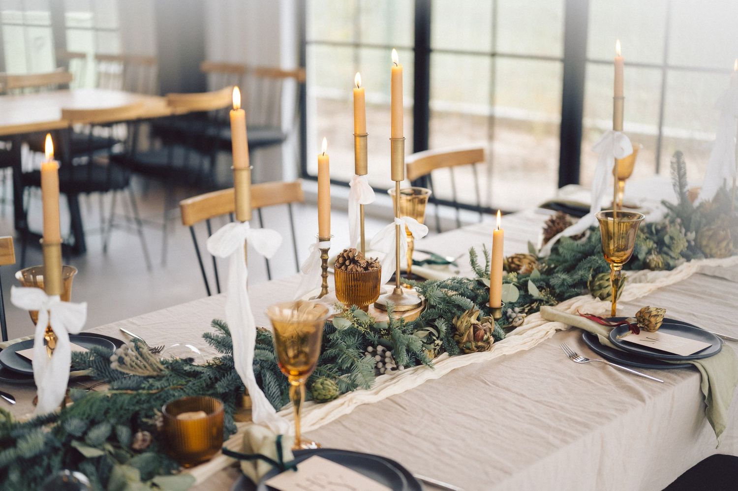 An elegant wedding reception table setting with gold candlesticks, greenery garlands and white table linens in a bright room.