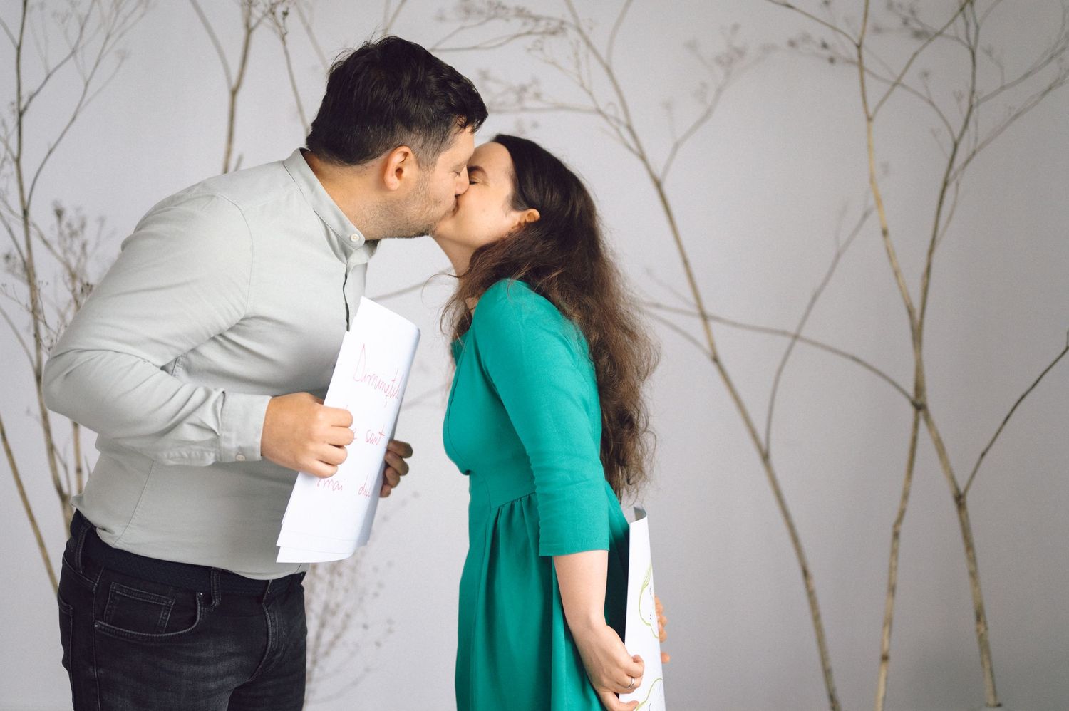 A couple in white and emerald green attire share a romantic kiss against a gray background with delicate branch patterns.