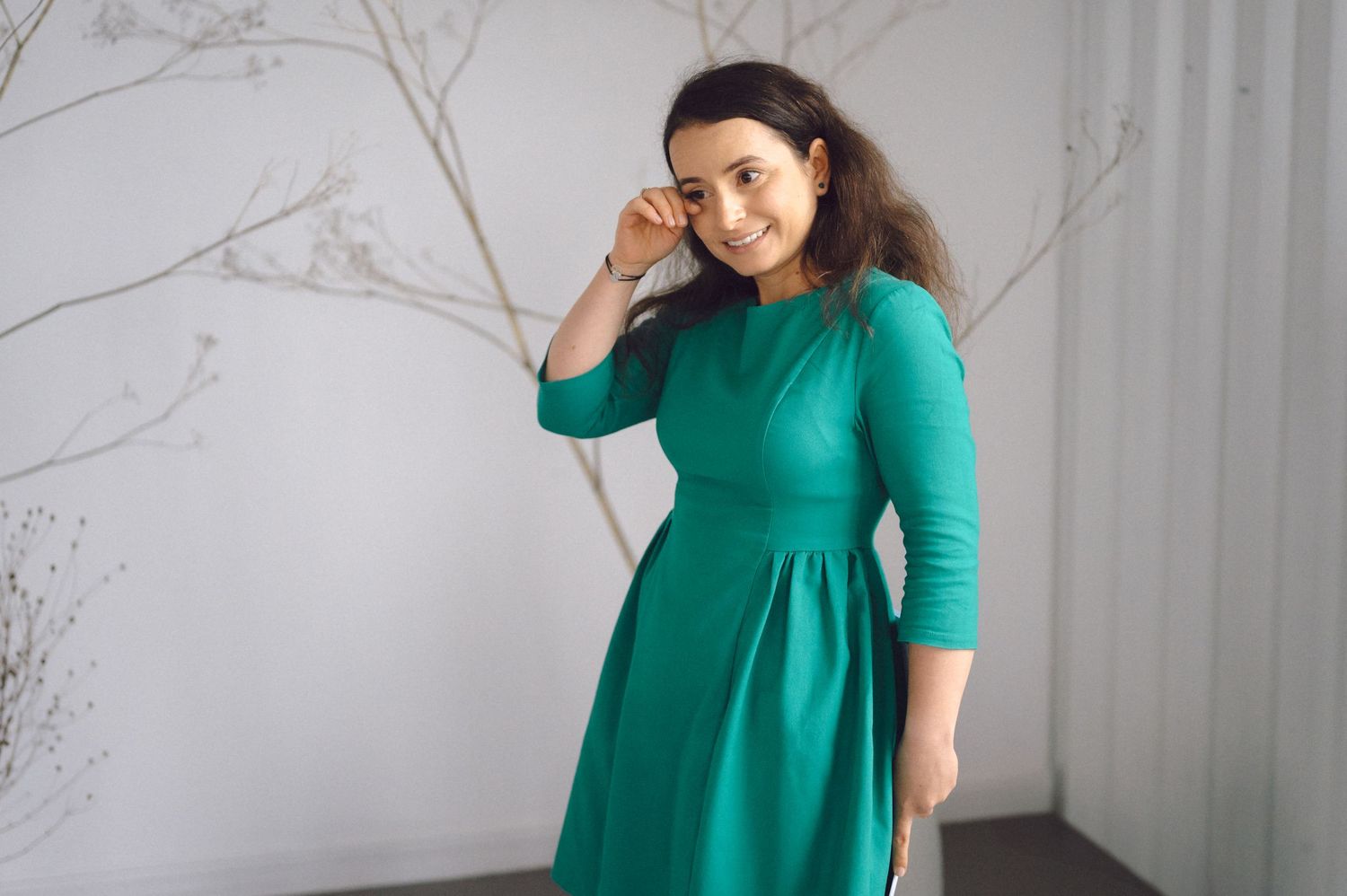 Someone wearing an elegant emerald green dress with three-quarter length sleeves poses against a light gray wall.
