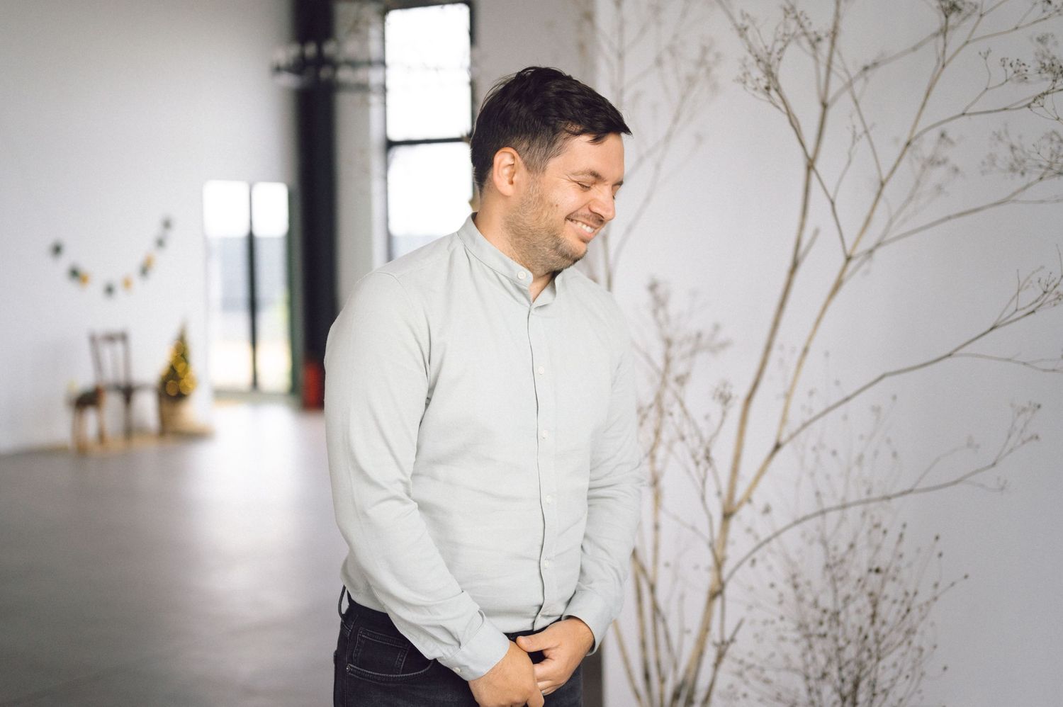 A person in a light gray sweater stands smiling by decorative branches in a bright minimalist interior space.