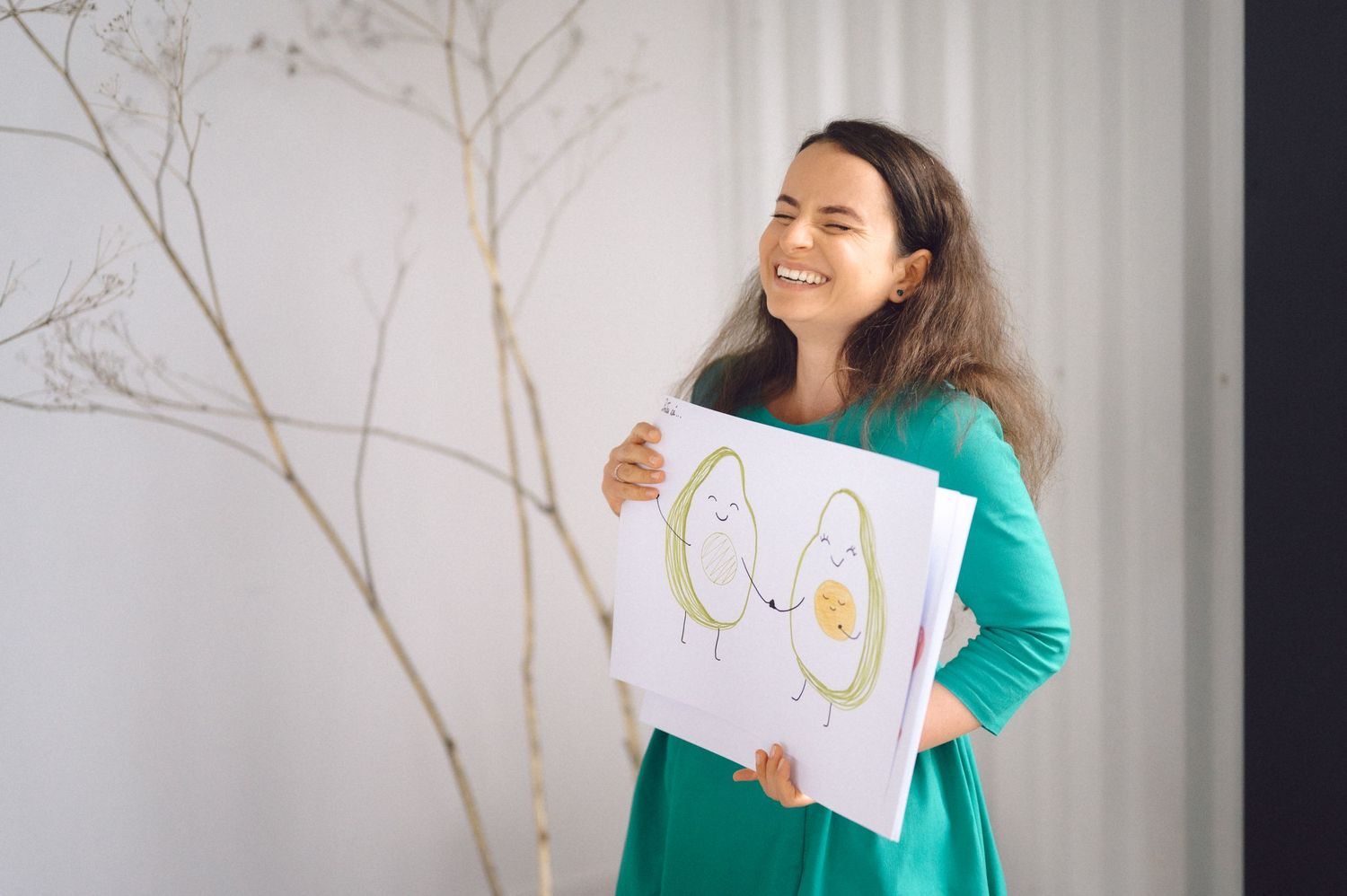 A woman in a teal dress holds up a handwritten sign while standing by decorative branches in a bright room.