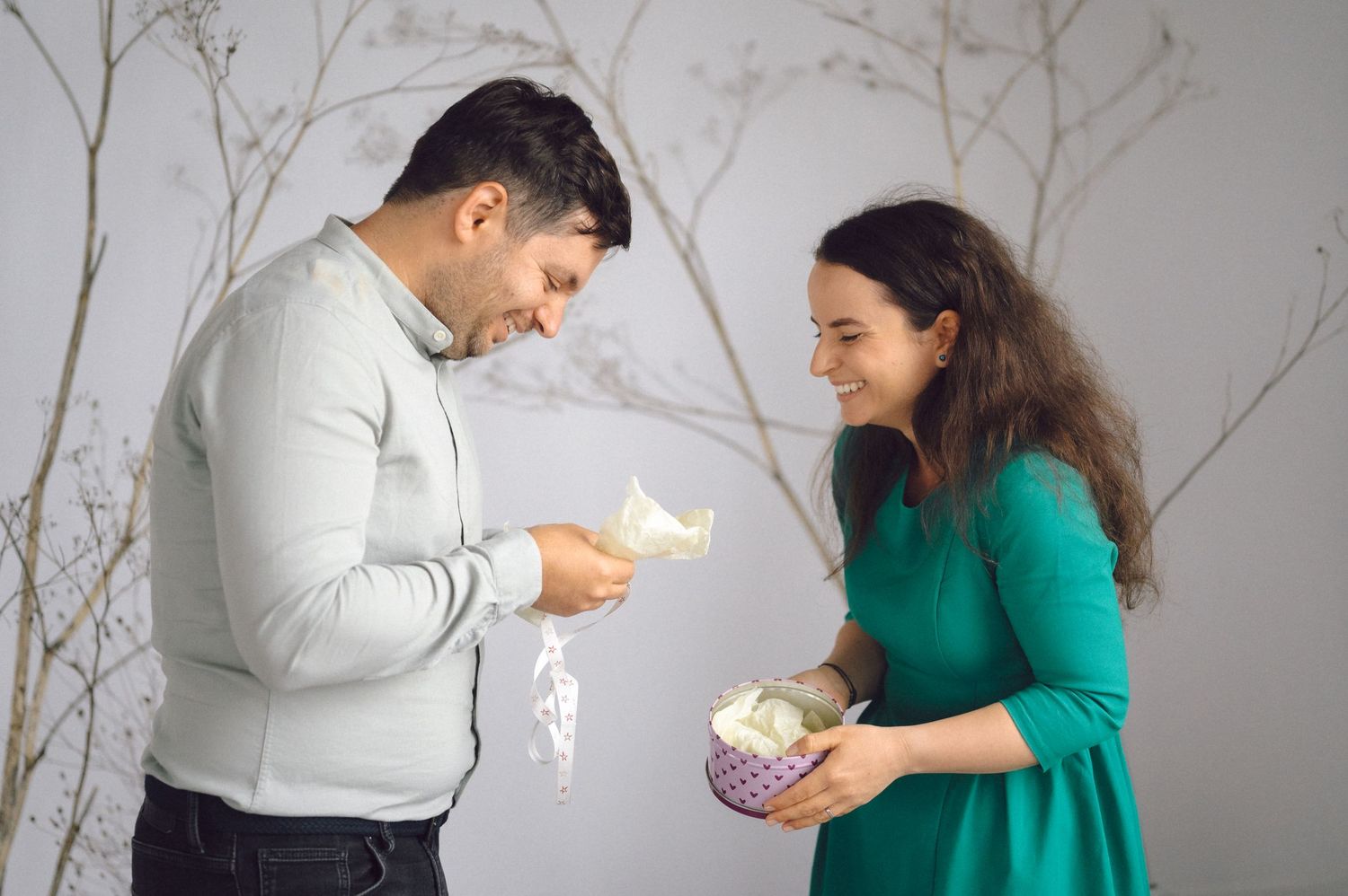 People in white and green sweaters share a humorous moment while exchanging gifts against a light gray background.