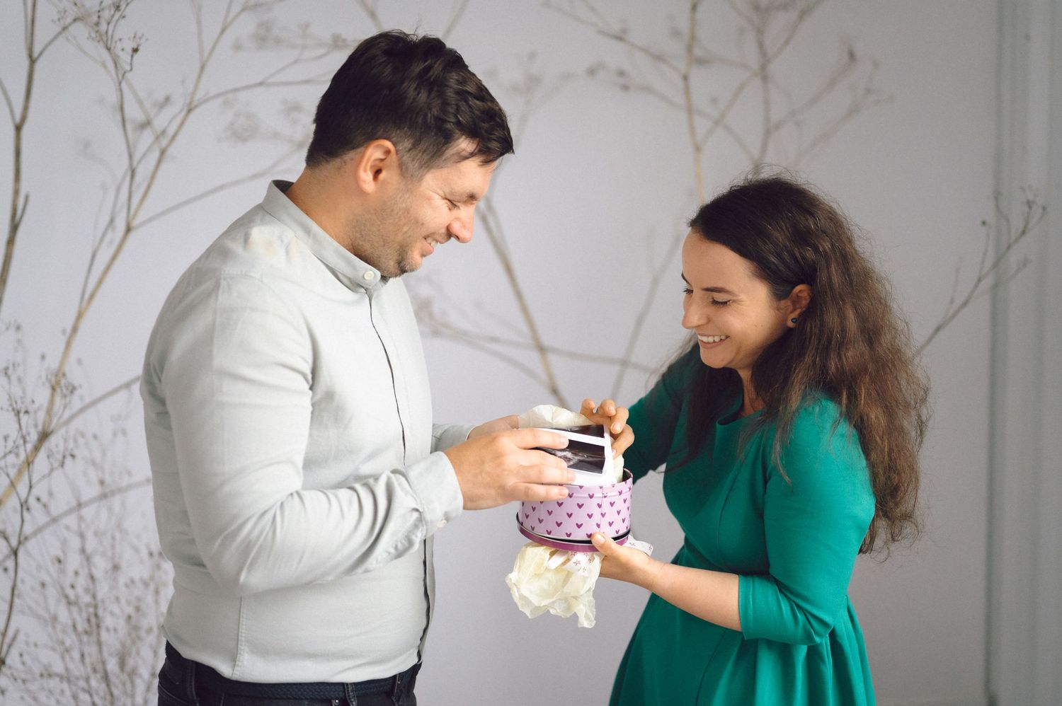 Two people sharing a happy moment while exchanging a small gift-wrapped present against a light gray wall.