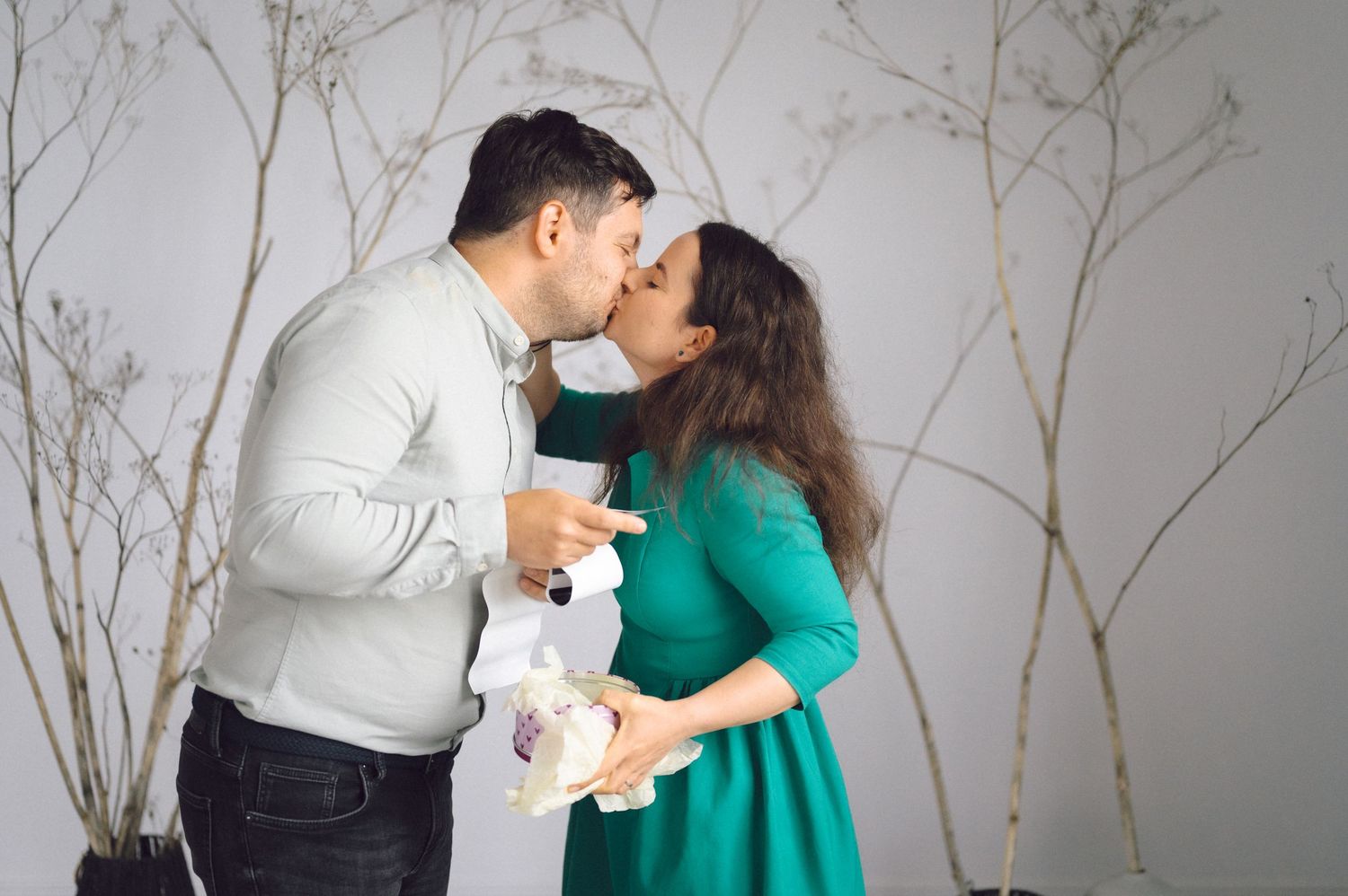 A couple in white and green attire share an intimate moment against a white wall with bare branches.