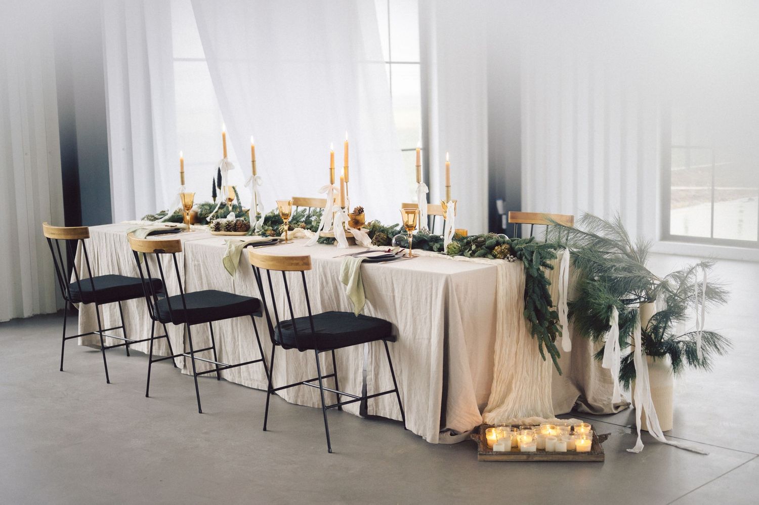 Modern wedding reception table with tall candles, eucalyptus greenery, and black metal chairs against white drapes.