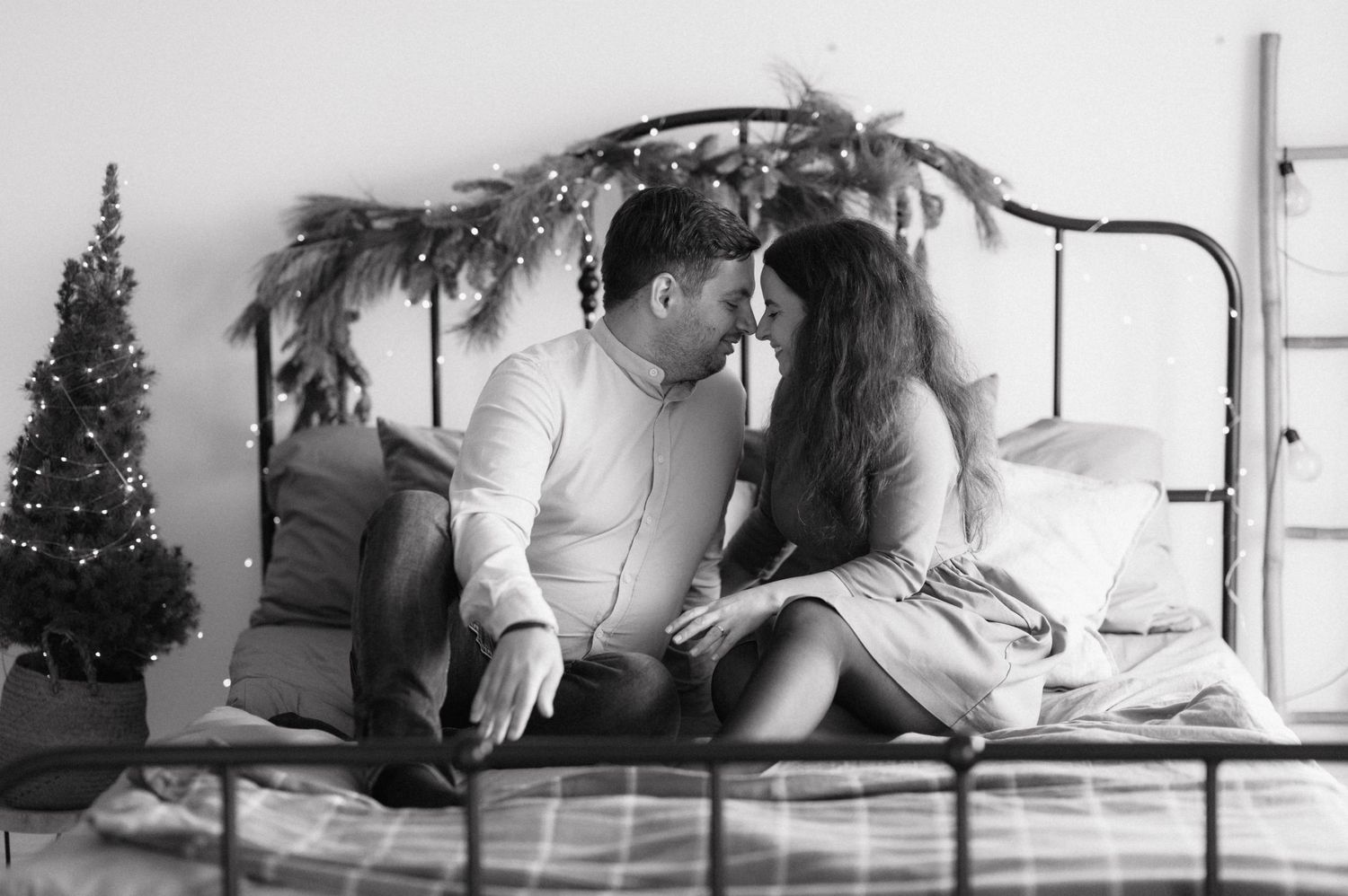 Couple shares intimate moment while sitting on bed with holiday decor and twinkling lights in black and white photograph.