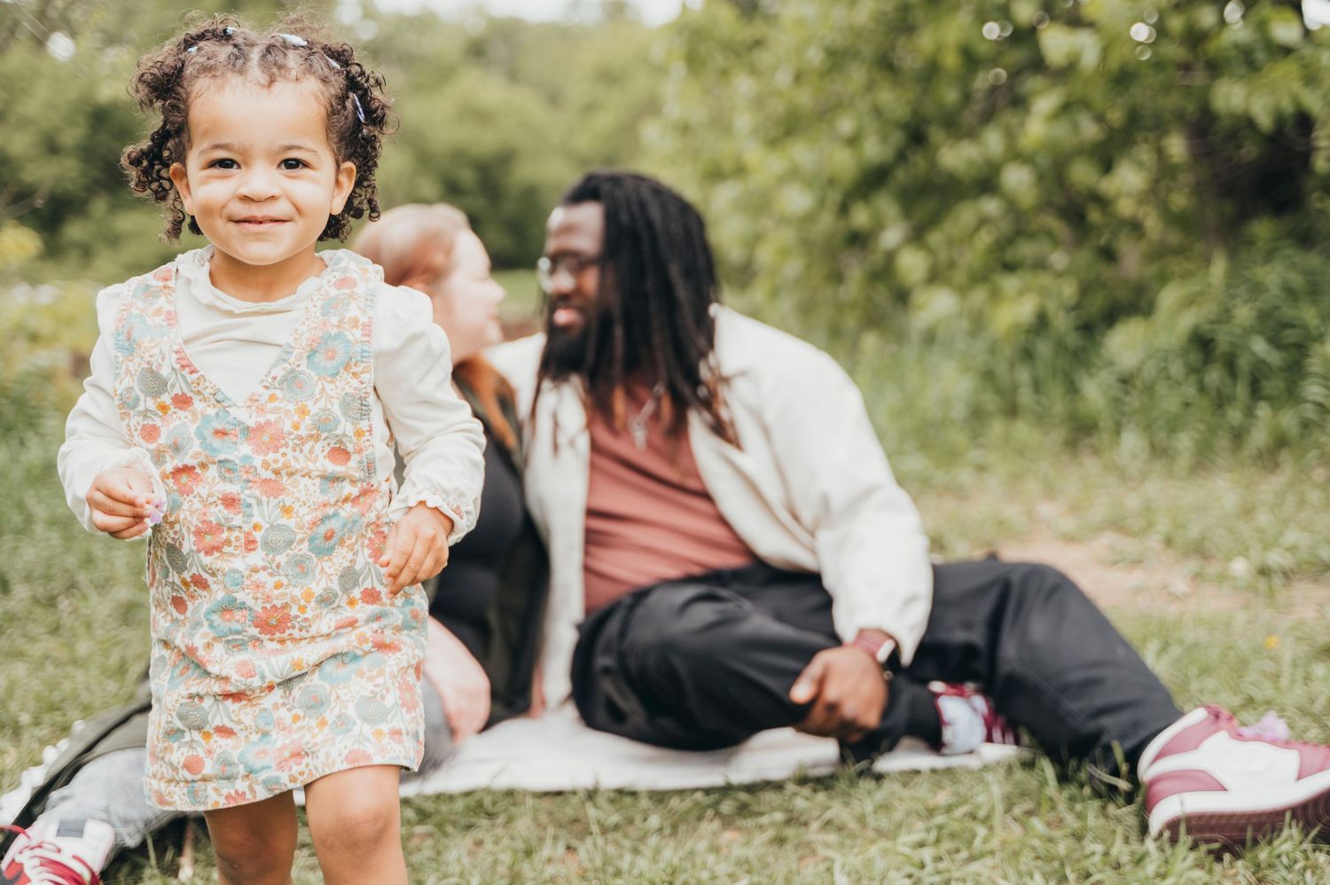 Cute toddler walks towards camera while parents look at each tore behind her in Mississauga park during the spring