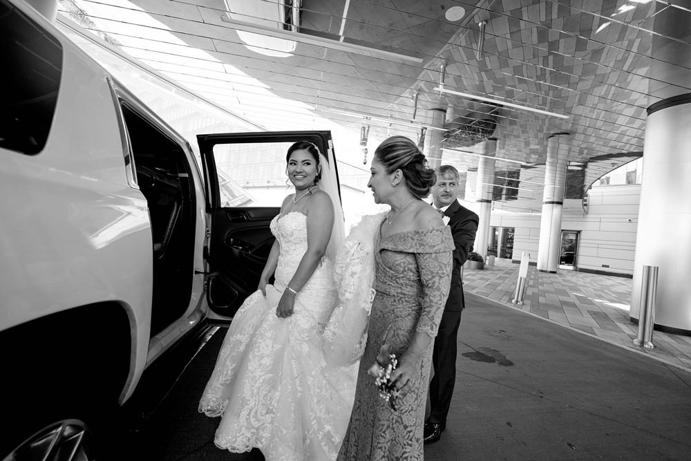 A bride in a white gown prepares to enter a limousine while accompanied by an attendant on a bright day.