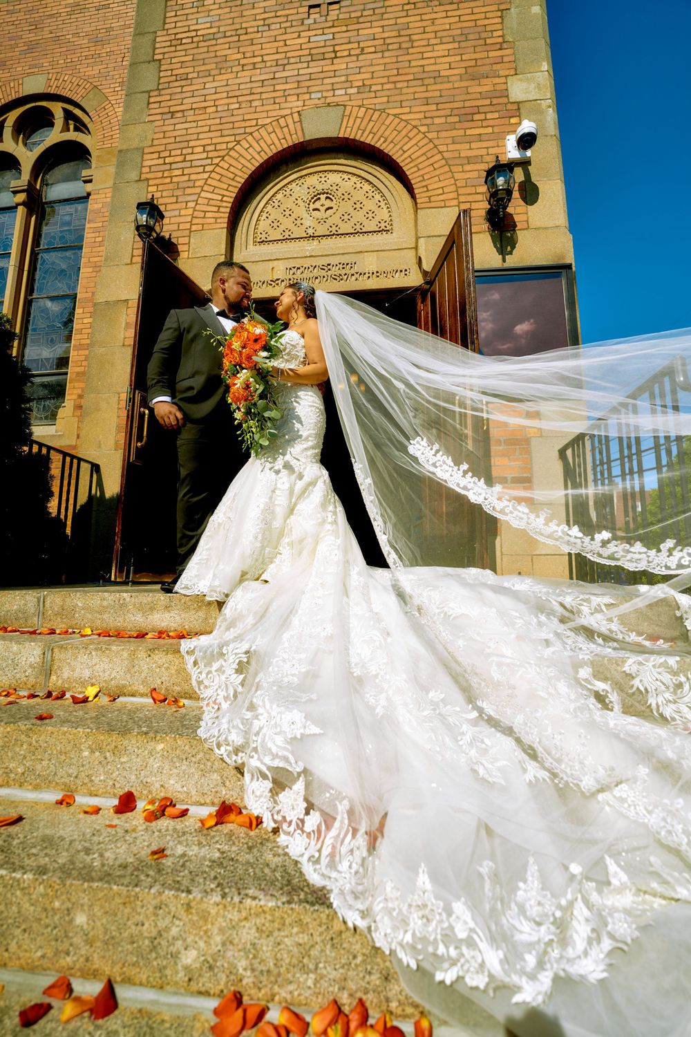 A dramatic wedding photo showcasing a flowing lace veil against historic brick church architecture.