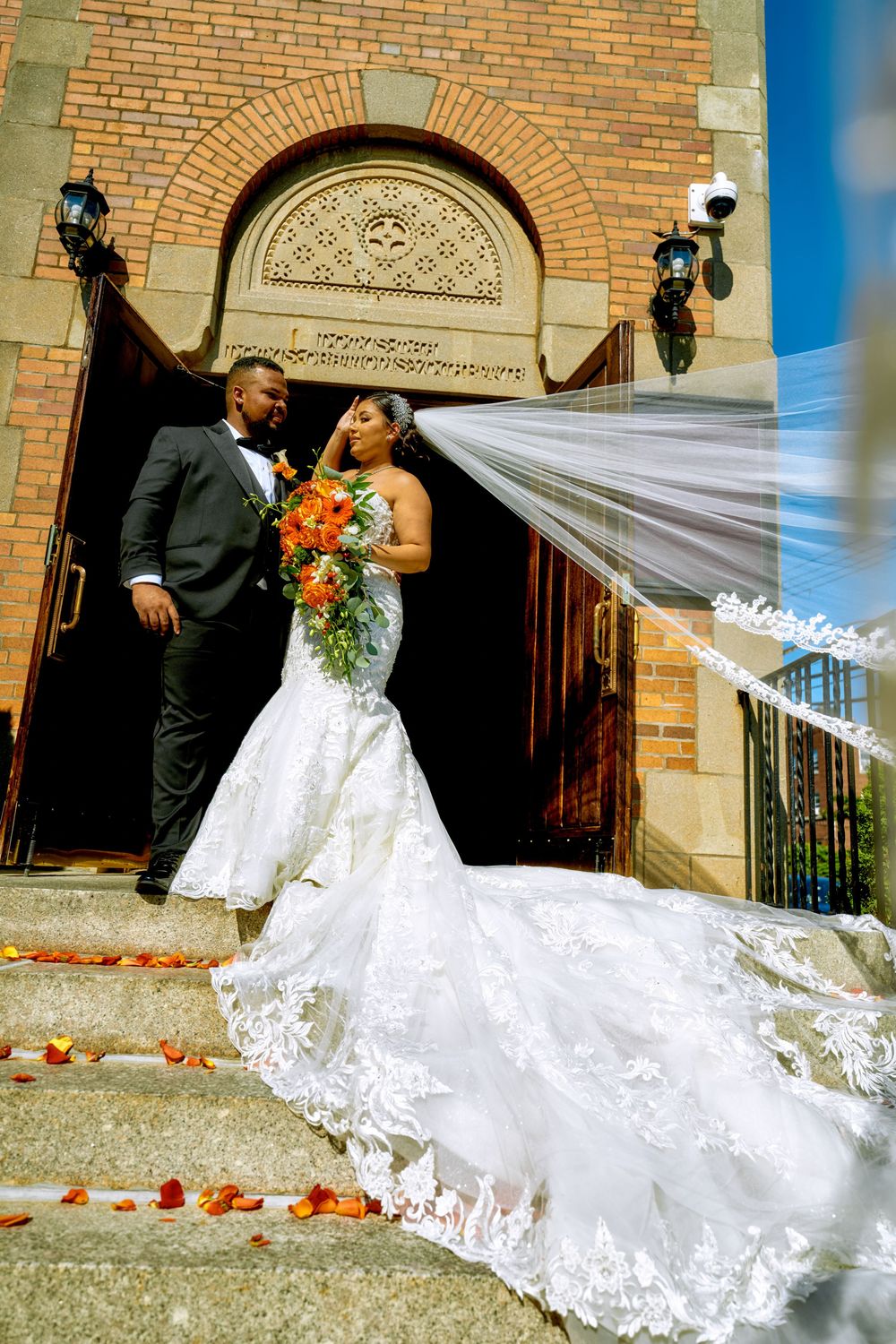A stunning bridal portrait with cascading veil and orange bouquet against brick church backdrop.