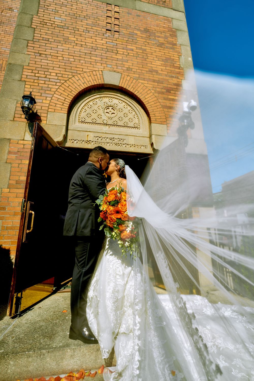A romantic moment captured on church steps with flowing veil and vibrant orange florals.