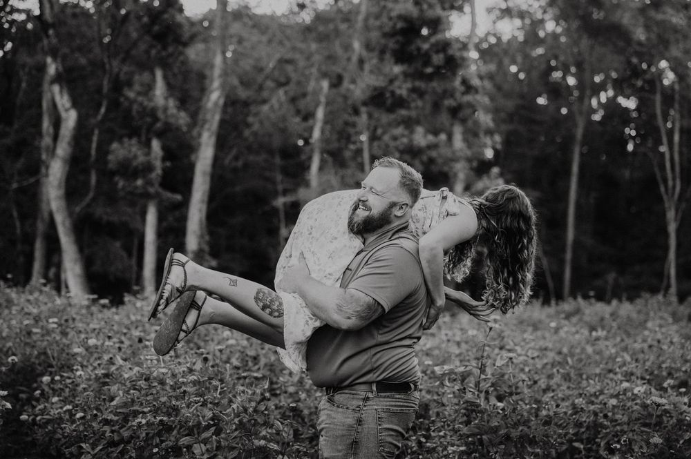 A romantic couple embraces and kisses in a lush garden setting with wildflowers and tall trees at sunset for their engagement photos