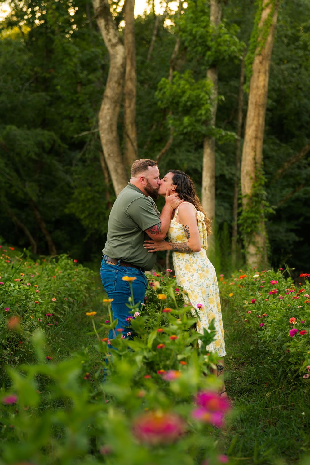 A romantic couple embraces and kisses in a lush garden setting with wildflowers and tall trees at sunset for their engagement photos