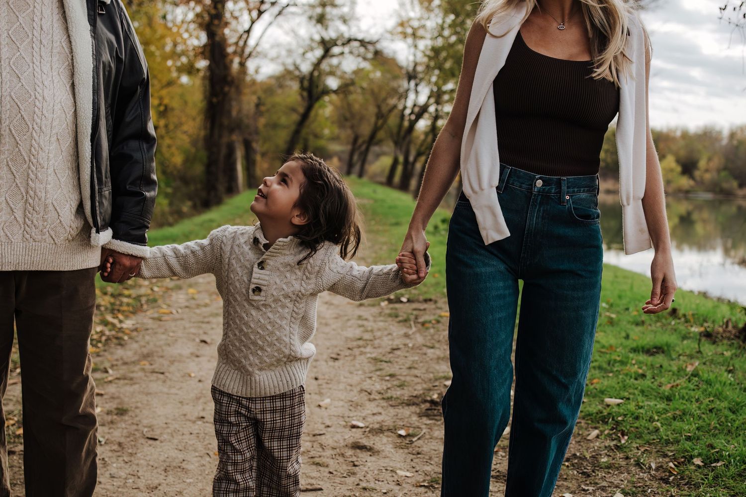 A small child in a knit sweater holds hands with adults while walking along a lakeside path. Fort Worth Fall Minis