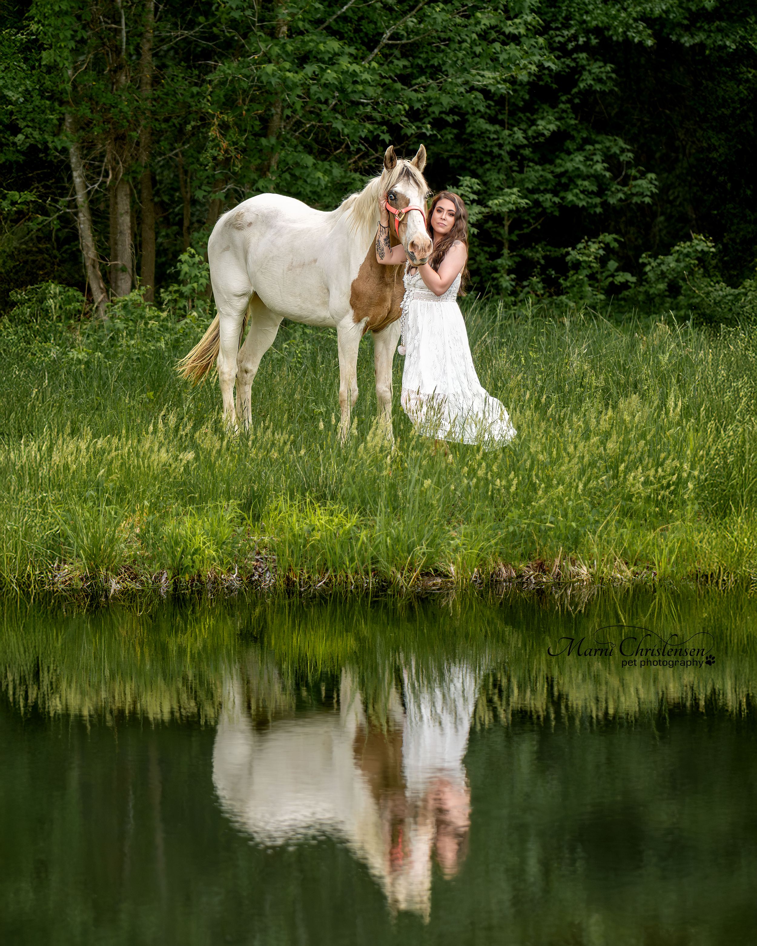A white horse and someone in a flowing white dress stand by a tranquil pond creating a mirror-like reflection.