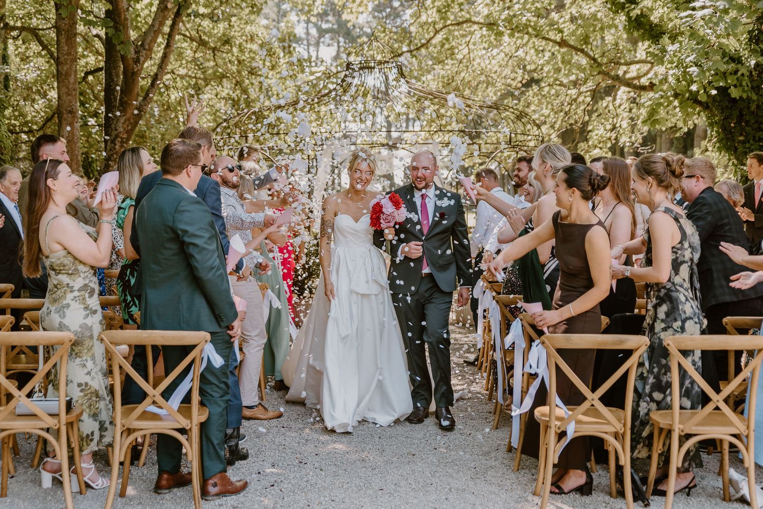 Wedding guests dance and celebrate during a vibrant outdoor reception surrounded by trees and string lights.