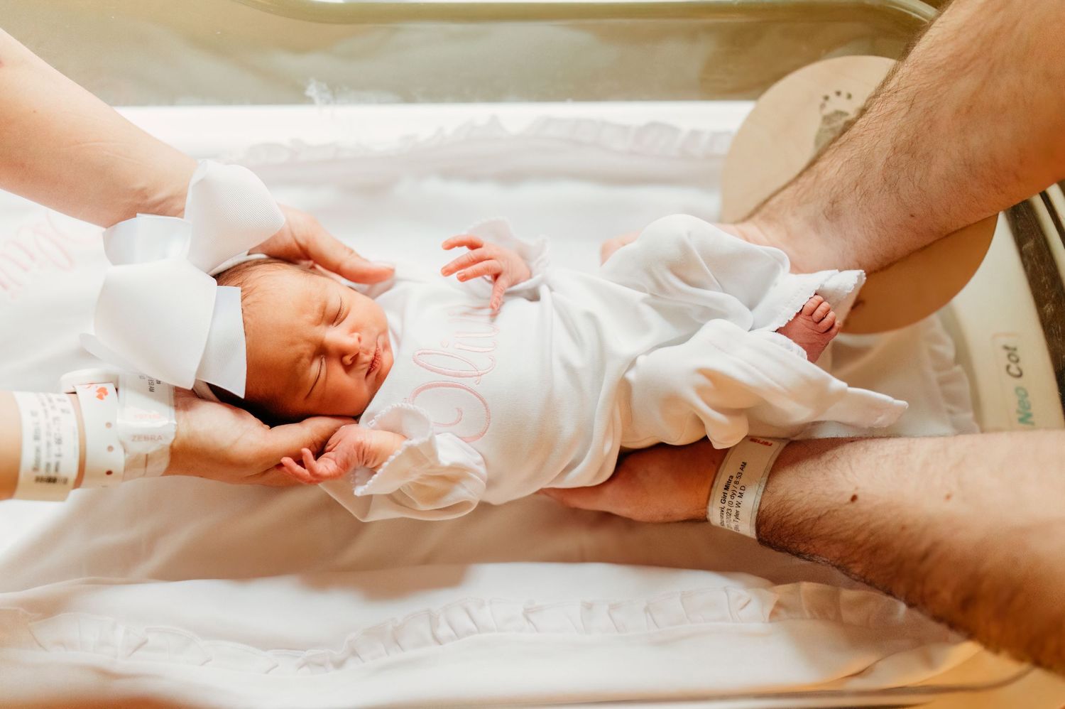 A newborn baby in white clothing lying on a soft surface in natural lighting.