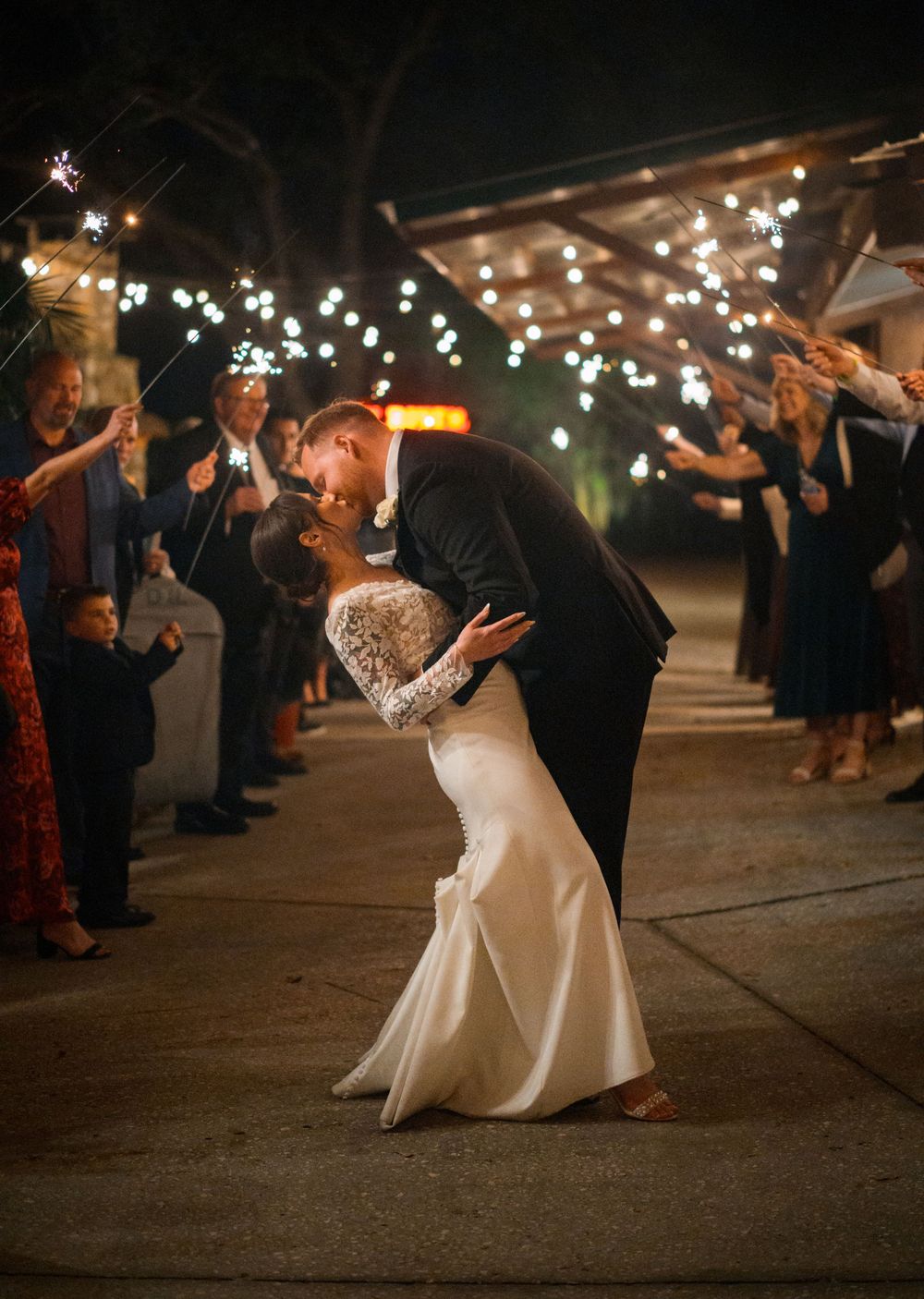 Romantic wedding couple shares a dip kiss under twinkling lights during their sparkler exit celebration.