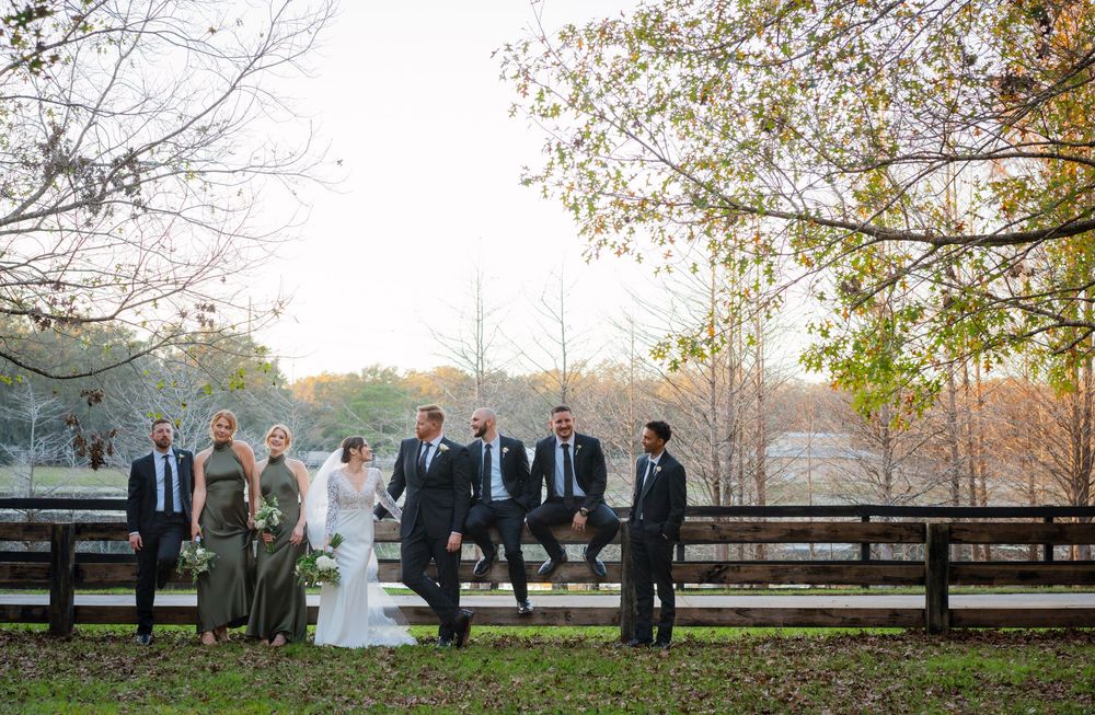 A wedding party stands by a rustic wooden fence at sunset with autumn trees in the background.