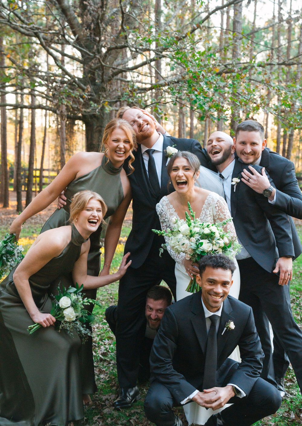 Wedding party in black attire poses playfully and energetically together in an outdoor wooded setting.
