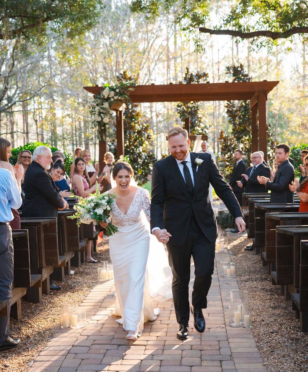 Newlyweds joyfully walk down a brick path between wooden pews during an outdoor woodland ceremony at sunset.