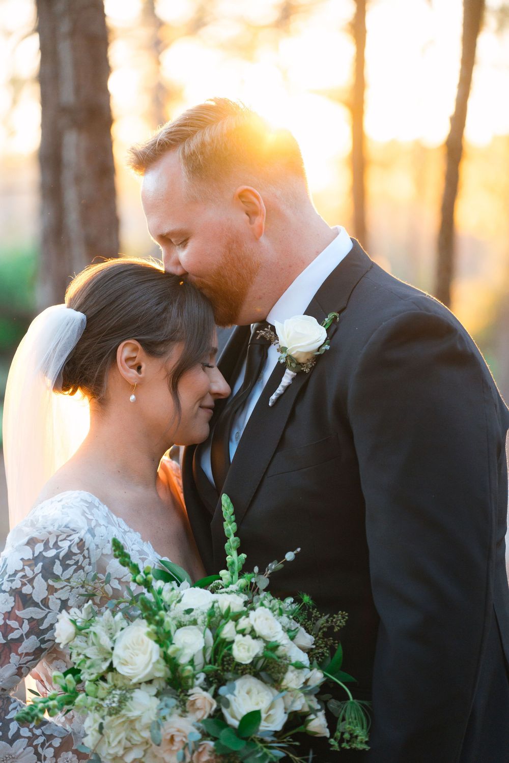 A romantic sunset wedding portrait shows a tender moment between newlyweds with a beautiful white floral bouquet.