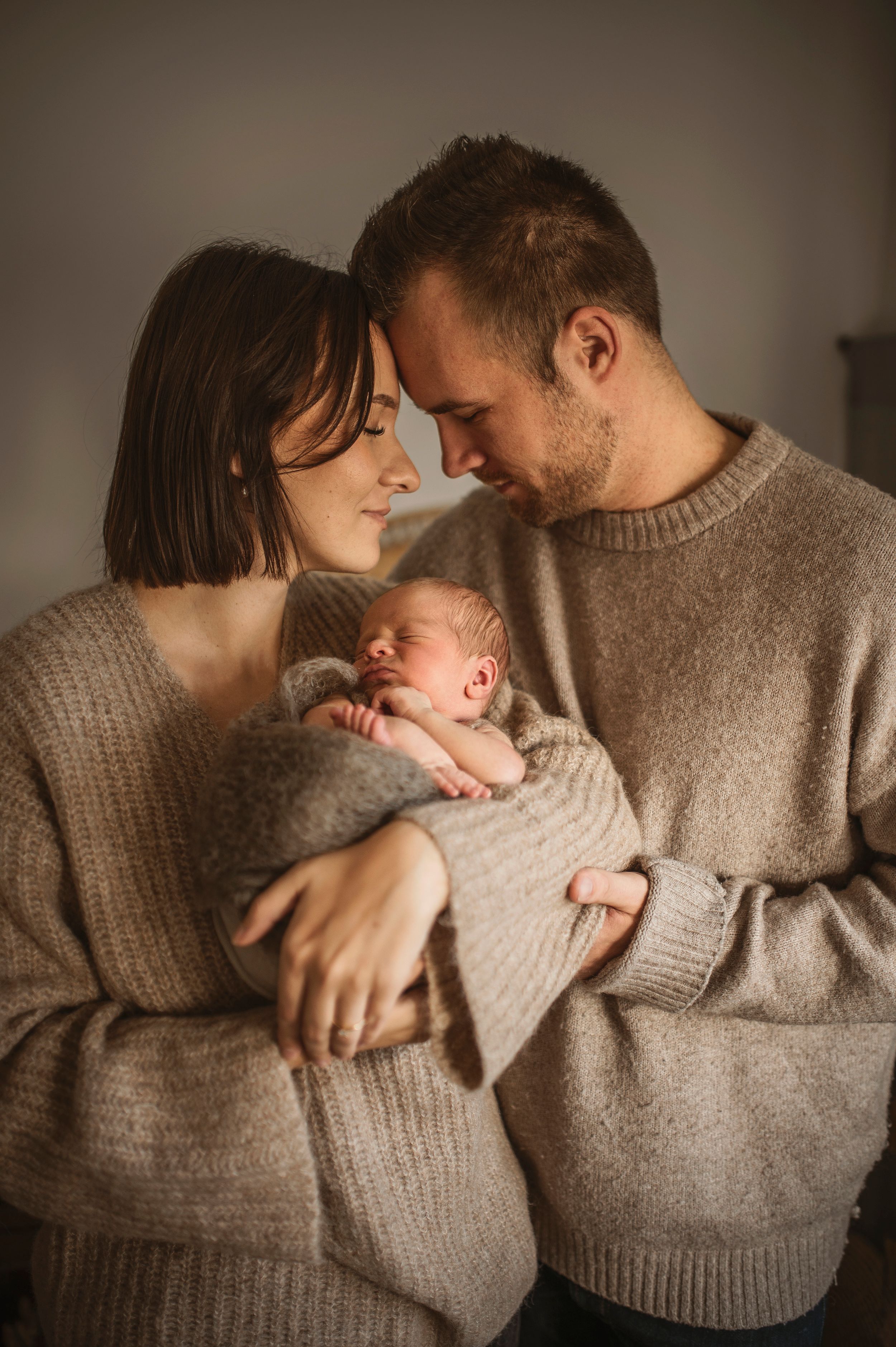 Family wearing matching beige sweaters cuddle with newborn baby in intimate lifestyle photoshoot.