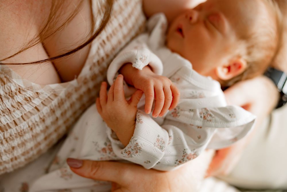 Newborn baby sleeps peacefully in white clothing while being gently held against a cozy knit sweater.