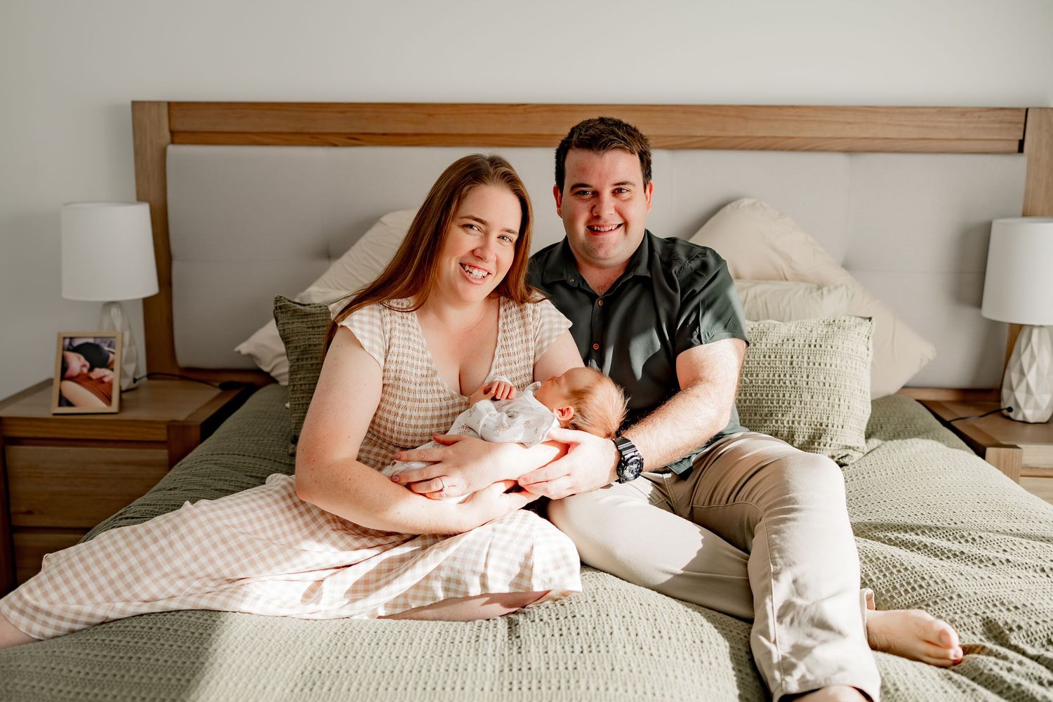 A couple sits on a bed and holds their newborn baby in a cozy bedroom setting.