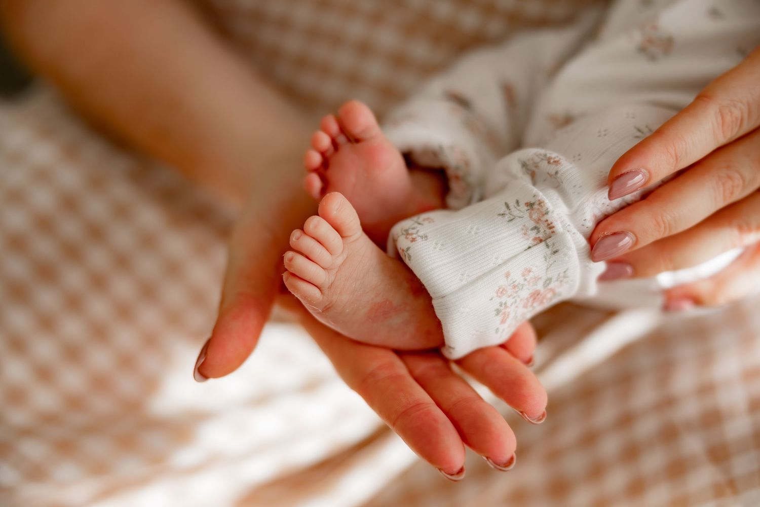 Adult hands gently dry tiny newborn baby feet with soft white towel on checkered surface.