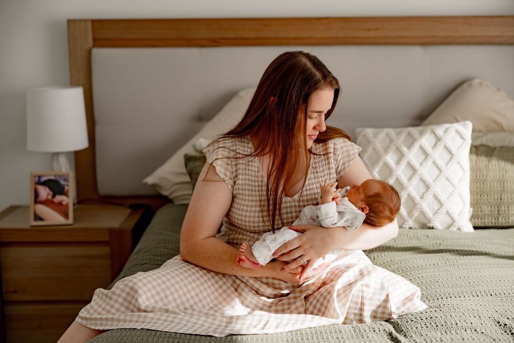 Mother in gingham dress sitting on bed tenderly holding and feeding newborn baby in cozy bedroom setting.