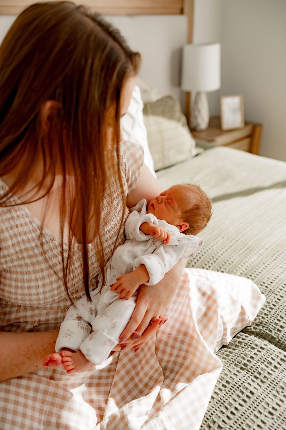 A person in gingham dress sits comfortably on a bed with soft white bedding and morning sunlight streaming in.