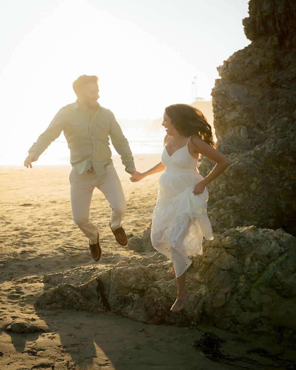 Couple in white outfits holding hands while exploring rocky beach formations at sunset.
