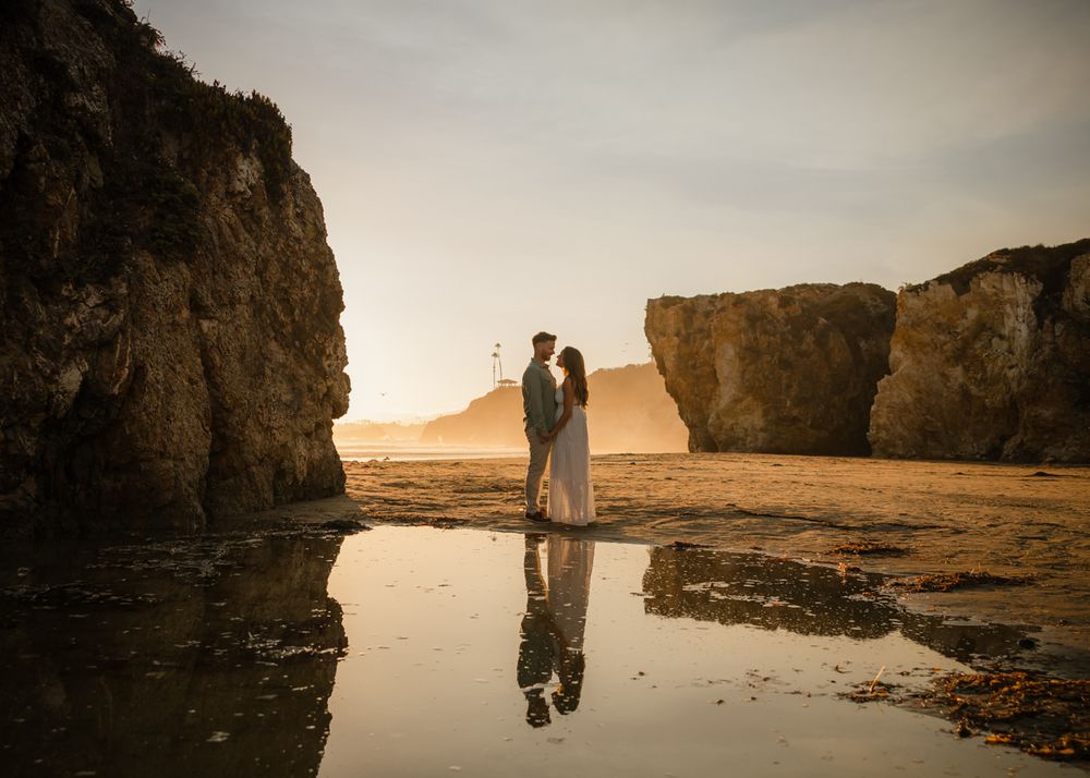 A silhouetted couple stands between dramatic coastal rock formations at sunset.
