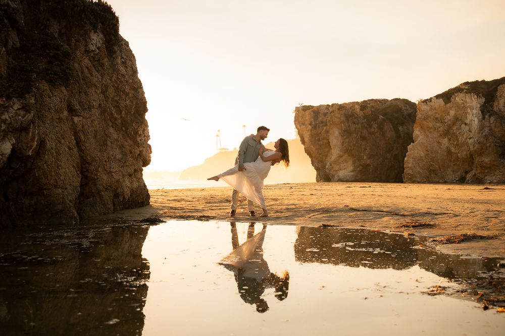 A romantic couple creates joyful poses between beach rock formations at sunset.