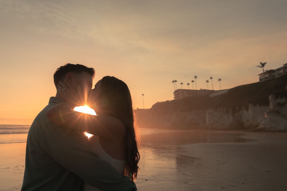 A backlit couple sharing an intimate moment on the beach during golden hour with cliffs in the distance.