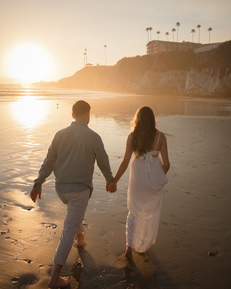 A couple walking hand in hand along a sunset beach with palm trees and cliffs in the background.