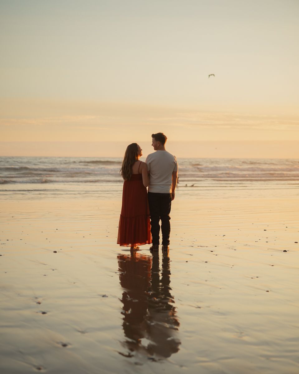 A couple stands silhouetted against a golden sunset on a reflective beach.