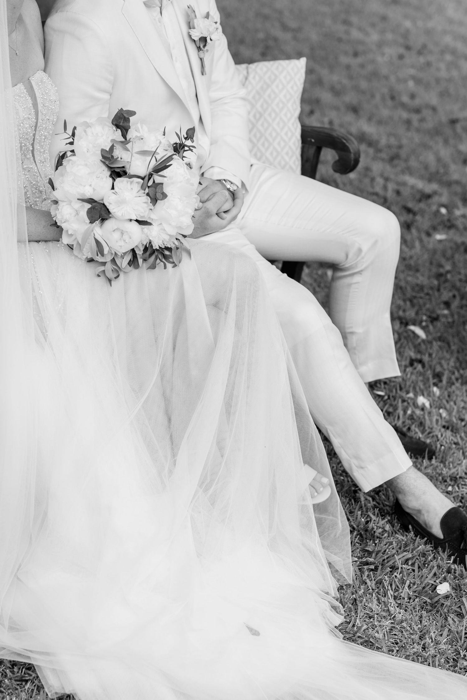 Ein Detailbild in Schwarz-Weiß von einem Hochzeitskleid, einem weißen Brautstrauß und dem Bräutigam im hellen Anzug, während ihrer Trauung.