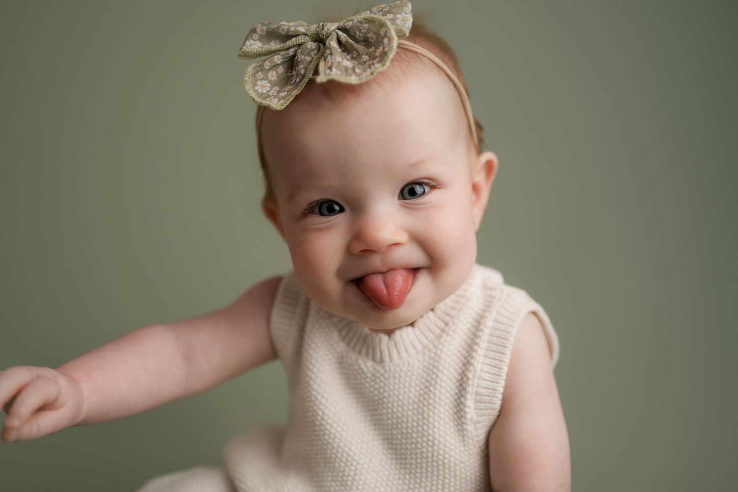 A cheerful baby girl in a white dress and bow sits smiling in a rattan chair on a dark background.