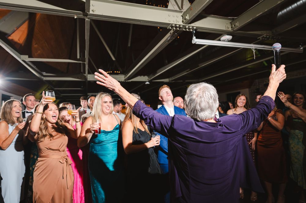 Grandmother singing to collected guests who are cheering in industrial reception hall with exposed beams at Drowned Lands Brewery Warwick NY