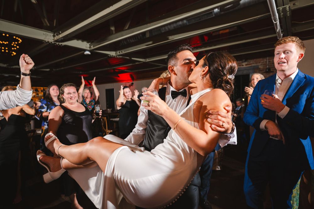 Groom lifting bride in celebration on dance floor during reception while guests dance at Drowned Lands Brewery in Warwick, NY Hudson Valley.