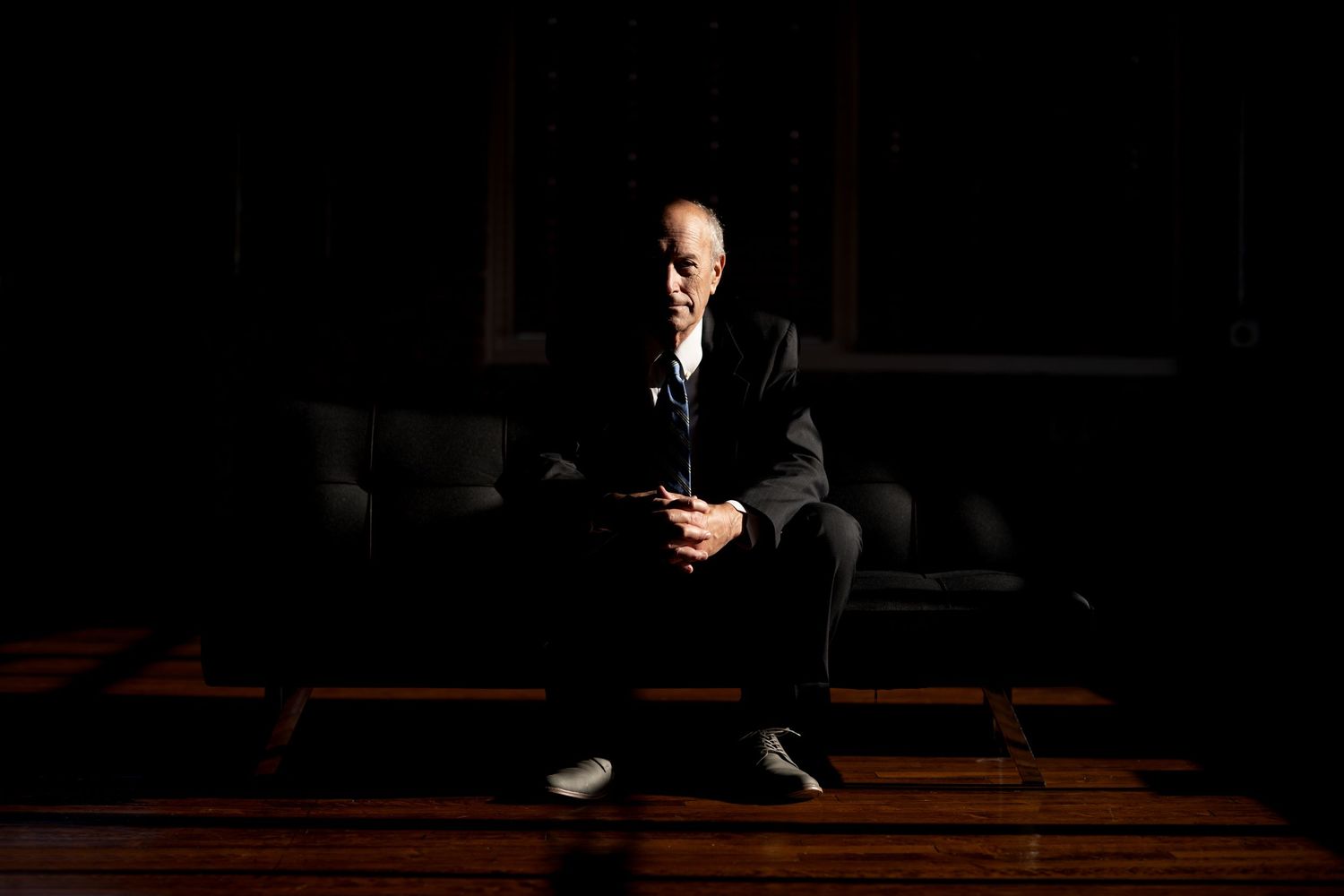Figure in a dark room sits thoughtfully on wooden stairs while dramatic lighting creates moody atmosphere.
