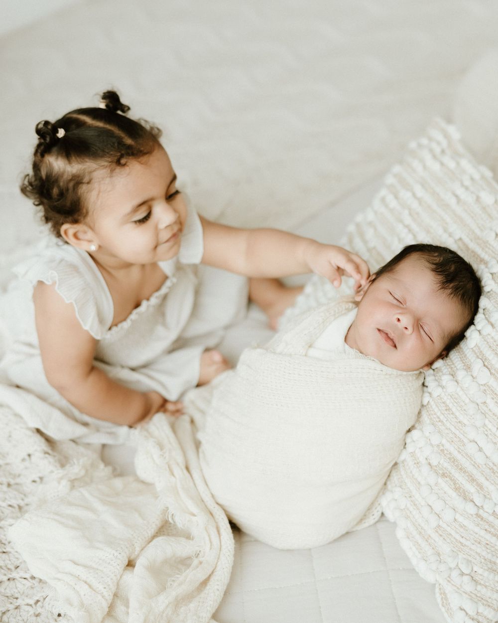 Two children in white clothing sit together on light-colored textiles in a sweet intimate moment.